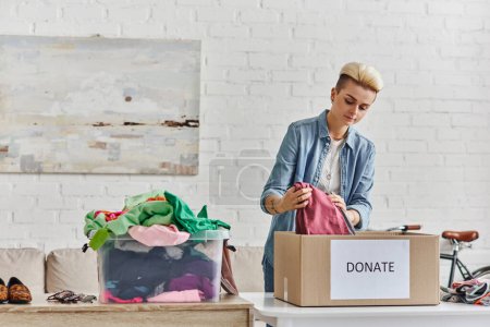 Spenden für einen guten Zweck, stylische tätowierte Frau verpackt Kleidungsstücke in Spendenbox in der Nähe von Plastikbehältern mit Second-Hand-Artikeln im Wohnzimmer, nachhaltiges Leben und soziales Verantwortungskonzept