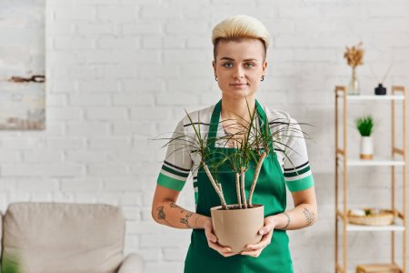 femme tatouée avec une coiffure à la mode, portant un tablier vert, tenant un pot de fleurs avec une plante tropicale et regardant la caméra dans l'appartement, respectueux de l'environnement, décor à la maison durable et concept de vie vert