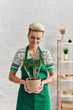überglückliche tätowierte Frau mit trendiger Frisur, die in grüner Schürze im modernen Wohnzimmer steht, Pflanzentherapie, nachhaltige Wohnkultur und grünes Wohnkonzept