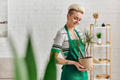 environmentally friendly habits, young and joyful tattooed woman in green apron holding potted exotic plant on blurred foreground in modern apartment, sustainable home decor and green living concept puzzle #661658328