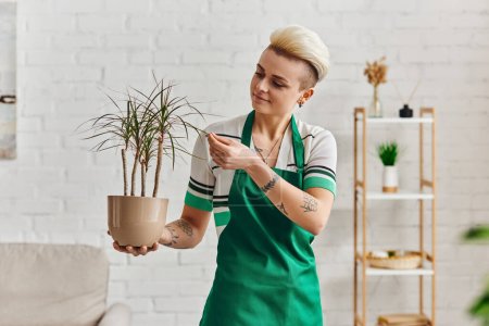 indoor gardening, young and positive tattooed woman in green apron touching leaves of tropical plant in modern apartment, plant care, sustainable home decor and green living concept mug #661658358