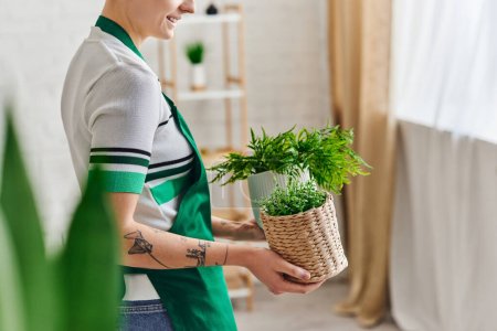 Téléchargez les photos : Vue partielle d'une jeune femme tatouée souriante dans un tablier tenant des pots de fleurs avec des plantes vertes dans le salon, un décor respectueux de l'environnement et durable et un concept de vie vert - en image libre de droit