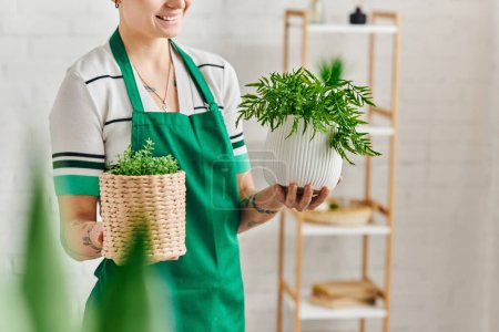 indoor gardening, partial view of happy tattooed woman in green apron holding flowerpots with houseplants in modern living room, blurred foreground, sustainable home decor and green living concept