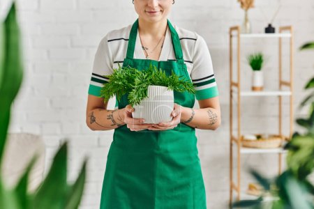 hogar sostenible, jardinería interior, vista parcial de la joven mujer tatuada en delantal sosteniendo maceta con planta verde y sonriendo en apartamento moderno, concepto de vida verde