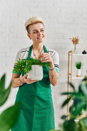 dreamy and happy tattooed woman in green apron looking away while standing with potted houseplant in modern living room, blurred foreground, sustainable home decor and green living concept