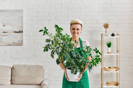 Pflanzenliebhaberin, stilvolle und aufgeregte Frau mit trendiger Frisur, die einen Blumentopf mit grüner Blattpflanze in der Hand hält und im Wohnzimmer in die Kamera blickt, nachhaltige Wohnkultur und grünes Wohnkonzept