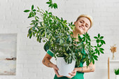eco-conscious lifestyle, positive and stylish tattooed woman with green foliage plant in flowerpot looking at camera in modern living room, sustainable home decor and green living concept magic mug #661658592