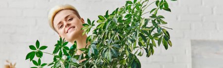 Porträt einer jungen Frau mit glücklichem Lächeln und trendiger Frisur, die in die Kamera schaut, in der Nähe grüner Zimmerpflanzen im Wohnzimmer, Indoor-Gärtnern, nachhaltiger Wohnkultur und grünem Wohnkonzept, Banner