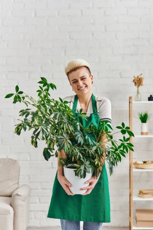 stilvolle und überglückliche Frau in grüner Schürze im modernen Wohnzimmer mit natürlicher Blattpflanze, umweltfreundlichen Gewohnheiten, nachhaltiger Wohnkultur und grünem Wohnkonzept