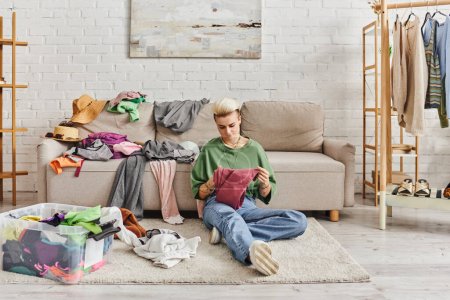 young tattooed woman in casual clothes sorting thrift store finds while sitting on floor near couch in modern living room, sustainable living and mindful consumerism concept