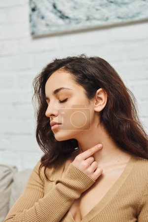 Junge brünette Frau in lässiger Kleidung, die Augen schließt, während sie eine Selbstmassage der Schilddrüse am Hals durchführt und den Lymphfluss zu Hause fördert, Selbstpflegungsritual und ganzheitliches Wellness-Praxiskonzept
