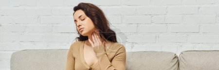 Young brunette woman in casual jumper doing massage for lymphatic system drainage and touching neck while sitting on couch at home, self-care ritual and holistic wellness practices concept, banner 