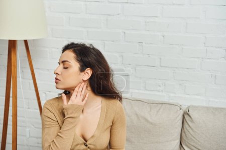 Téléchargez les photos : Vue latérale de la jeune femme brune en pull brun massant le cou pendant le massage lymphatique à domicile et assise sur le canapé, concept de guérison holistique et rituel d'autosoin, soulagement de la tension - en image libre de droit