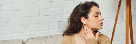 Photo for Side view of young brunette woman in casual brown jumper massaging lymphatic nodes on neck during self-massage on couch at home, self-care ritual and holistic healing concept, banner, tension relief - Royalty Free Image