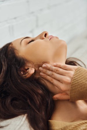 Young brunette woman in brown jumper touching lymphatic node on neck and relaxing during self-massage on couch at home, self-care ritual and holistic healing concept, tension relief