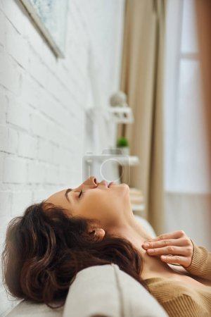 Photo for Side view of young brunette woman in brown jumper massaging thyroid gland and neck while relaxing and sitting on couch at home, self-care ritual and holistic healing concept, tension relief - Royalty Free Image