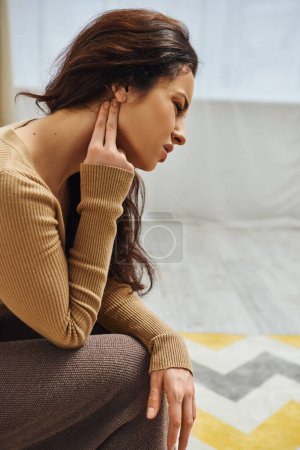 Photo for Side view of displeased young brunette woman in casual clothes touching neck during self-massage in blurred house, self-care ritual and holistic healing concept, tension relief - Royalty Free Image