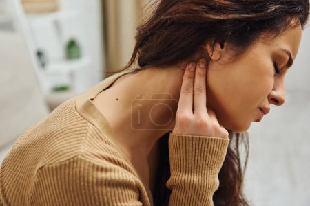 Photo for Side view of young brunette woman suffering from pain while doing lymphatic self-massage in blurred house, self-care ritual and holistic healing concept, tension relief, balancing energy - Royalty Free Image