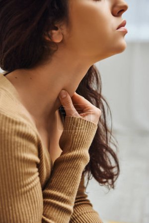 Widok na brunetkę młodą kobietę w brązowym swetrze masującym Tyrode 'a na szyi podczas masażu w domu, rytuał samoopieki i holistyczna koncepcja uzdrawiania, równoważenie energii