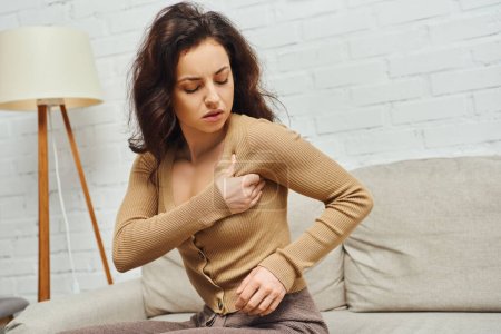 Brunette woman in casual brown jumper massaging lymphatic nodes on armpit while doing drainage and sitting on couch at home, enhancing self-awareness and body relaxation concept, balancing energy