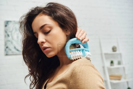 Portrait of young brunette woman in casual jumper holding handled massager and doing shoulder self-massage in blurred house, enhancing self-awareness and body relaxation concept, balancing energy