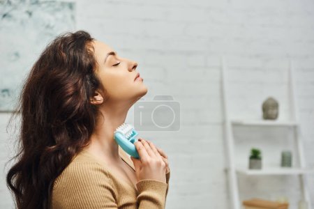 Widok z boku relaksującej młodej brunetki w brązowym swetrze masażu tarczycy i układu limfatycznego z obsługiwanym masażerem w domu, zwiększenie samoświadomości i koncepcji relaksu ciała