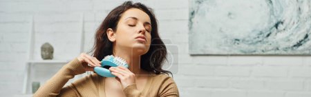 Téléchargez les photos : Jeune femme brune détendue en pull casual massant le cou avec masseur lors de l'auto-massage du système lymphatique dans le salon, rituel d'auto-soin et concept holistique de bien-être et de santé, bannière - en image libre de droit