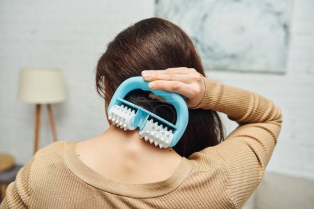 Back view of brunette woman massaging neck and back with modern handled massager while standing in blurred living room, lymphatic system support and home-based massage, tension relief