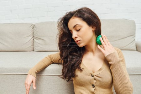 Young brunette woman in brown jumper massaging neck with manual massage ball while sitting near couch in living room, lymphatic system support and home-based massage, tension relief
