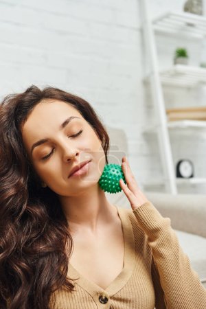 Portret zadowolonej brunetki z zamkniętymi oczami w brązowym swetrze masującym węzły chłonne z ręczną piłką do masażu w salonie, wsparciem układu limfatycznego i masażem domowym