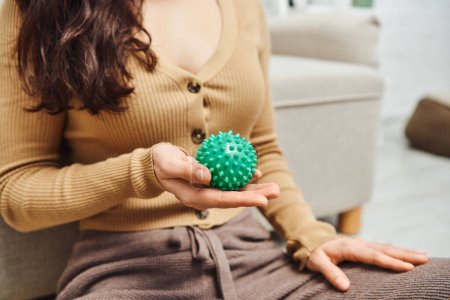 napięcie, przycięty widok niewyraźne brunetka kobieta w ubraniu casual trzymając ręczny masaż piłkę siedząc w pobliżu kanapy w domu, wsparcie układu limfatycznego i masażu domowego 