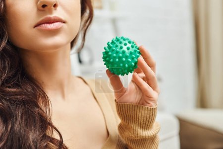 Widok upraw młodej brunetki w swetrze trzymając ręczną piłkę do masażu stojąc w rozmytym salonie w domu, wsparcie układu limfatycznego i masaż domowy, rozładowanie napięcia