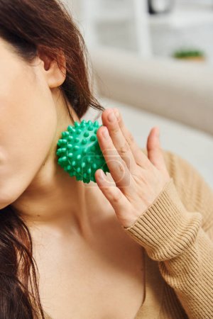 Cropped view of young woman in brown jumper massaging lymphatic nodes with manual massage ball for drainage at home, lymphatic system support and home-based massage, tension relief