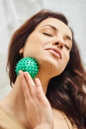 Portret zadowolonej młodej brunetki z zamkniętymi oczami masująca szyję z ręczną piłką do masażu w domu, wsparcie układu limfatycznego i masaż domowy, równoważąca energia