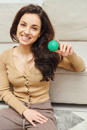 Portret wesołej brunetki w luźnych ubraniach uśmiechniętej do aparatu i trzymającej ręczną piłkę do masażu w pobliżu kanapy w domu, masaż domowy i holistyczna koncepcja praktyk odnowy biologicznej, równoważąca energię