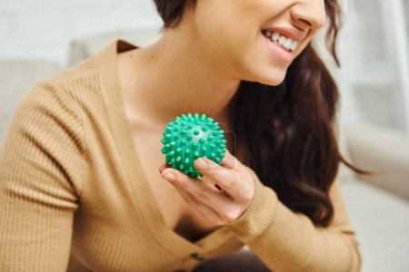 Photo for Cropped view of cheerful young brunette woman in brown jumper holding manual massage ball in blurred living room, body relaxation and holistic wellness practices, balancing energy - Royalty Free Image