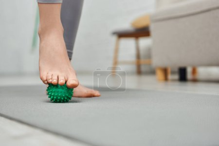 Widok kobiety masującej stopę ręczną piłką do masażu i stojącej na macie fitness w domu, relaksacja ciała i holistyczne praktyki odnowy biologicznej, równoważenie energii
