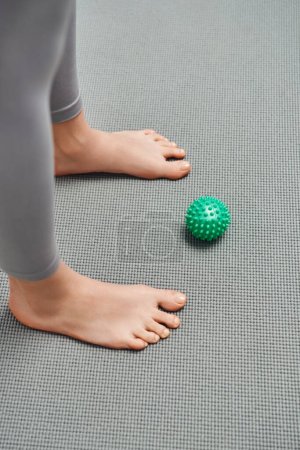 Vista superior de la bola de masaje manual cerca de la mujer descalza de pie en la estera de fitness en el hogar, relajación corporal y prácticas de bienestar holístico, energía de equilibrio
