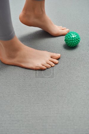 Ausgeschnittene Ansicht einer barfüßigen Frau, die auf einer Fitnessmatte in der Nähe eines manuellen Massageballs zu Hause steht, Körperentspannung und ganzheitliche Wellnesspraktiken, die Energie ausgleichen