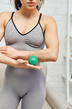 Ausgeschnittene Ansicht einer jungen Frau in Sportkleidung, die einen manuellen Massageball in der Hand hält und im verschwommenen Wohnzimmer steht, ausgleichende Energie und ganzheitliches Heilkonzept, myofasziale Befreiung
