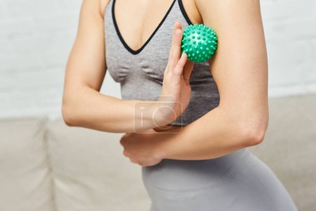 Widok upraw kobiety w odzieży sportowej masujący mięśnie na ramieniu z ręczną piłką do masażu w rozmytym salonie, równoważąca energia i całościowa koncepcja gojenia, uwalnianie mięśnia sercowego