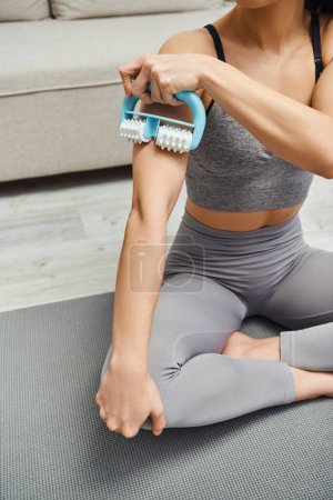 Widok upraw młodej kobiety w odzieży sportowej masaż mięśni na ramieniu z obsługiwanym masażerem podczas siedzenia na macie fitness w domu, równoważenie energii i holistycznej koncepcji gojenia, uwolnienie mięśnia sercowego