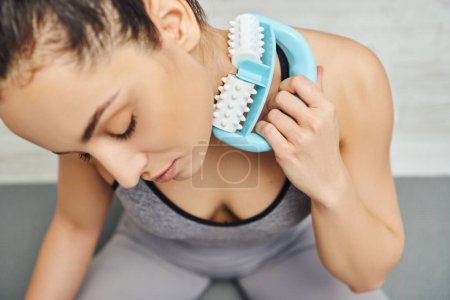 Overhead view of relaxed woman in sportswear massaging neck with modern handled massager and sitting on blurred fitness mat at home, home-based massage and holistic wellness practices concept