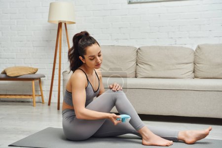 Young brunette and barefoot woman in activewear using handled massager on leg while sitting on fitness mat near couch in living room at home, inner peace and harmony concept, myofascial release