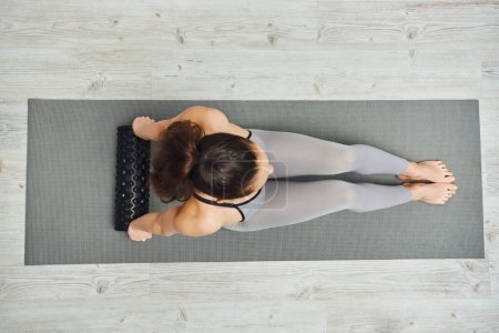 Overhead view of young brunette woman in activewear holding roller massager while sitting on fitness mat at home, home-based massage and holistic practices concept, myofascial release
