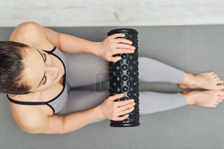 Widok na młodą brunetkę w odzieży sportowej trzymającą masażer rolkowy podczas siedzenia na macie fitness w domu, masaż domowy i koncepcja holistycznych praktyk, uwolnienie mięśnia sercowego