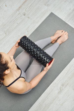 Overhead view of young barefoot woman in activewear holding roller massager and sitting on fitness mat in living room, home-based massage and holistic practices concept, myofascial release