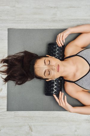 Widok z góry na młodą, zrelaksowaną brunetkę w odzieży sportowej z masażerem rolkowym na szyi leżącą na macie fitness w domu, masażem domowym i koncepcją holistycznych praktyk, uwalnianiem mięśnia sercowego