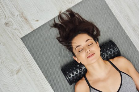 Vista superior de la joven morena relajada con los ojos cerrados usando masajeador de rodillos en el cuello mientras está acostado en la alfombra de fitness en casa, masaje basado en el hogar y el concepto de prácticas holísticas, liberación miofascial