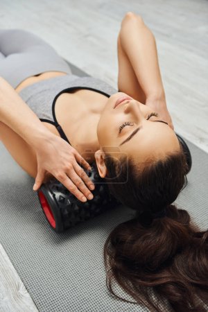 Young brunette woman in activewear massaging neck with roller massager and relaxing on fitness mat in living room at home, focus on self-care and well-being concept, tension relief